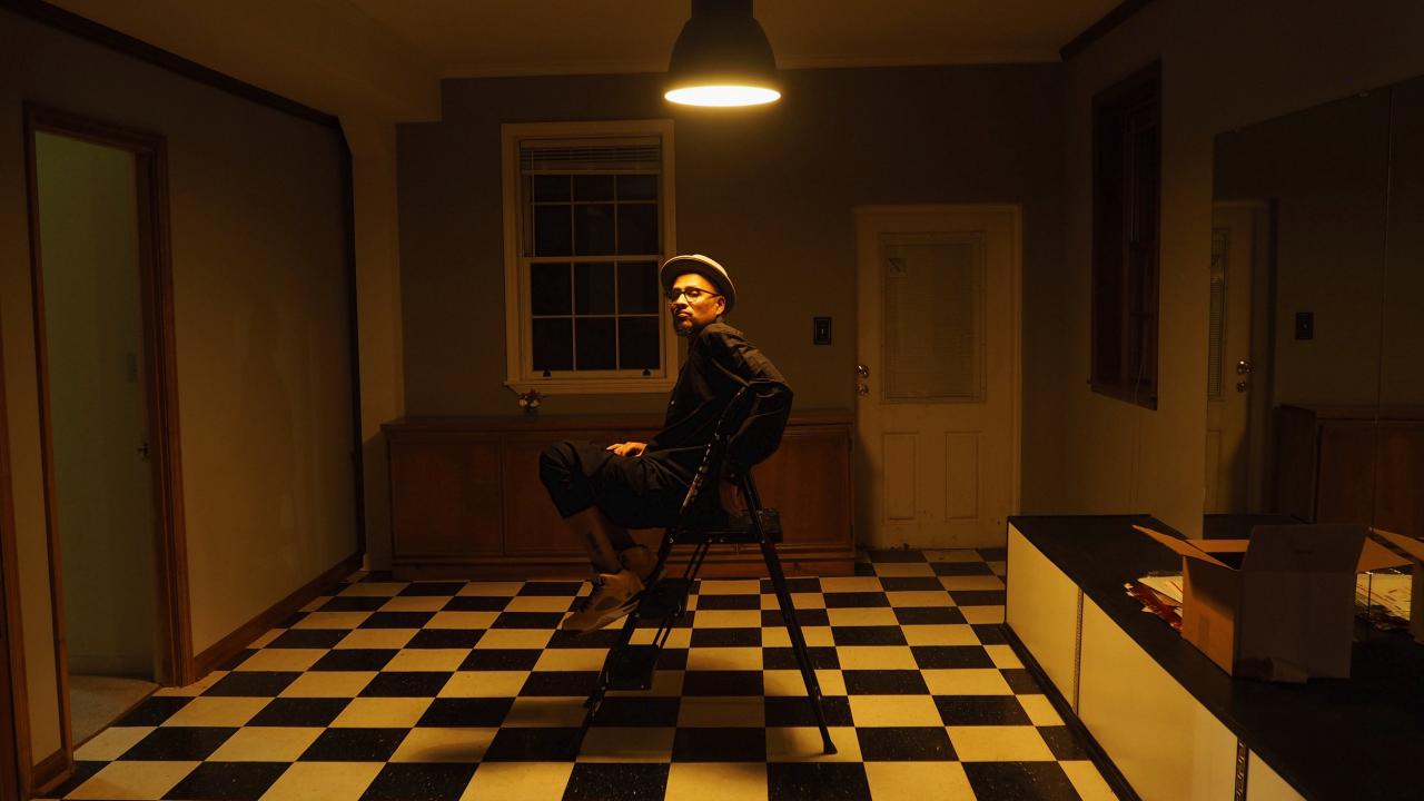 A photo of a man sitting in a room bathed in warm light. He looks at the viewer under one ceiling lamp. A checkered floor.