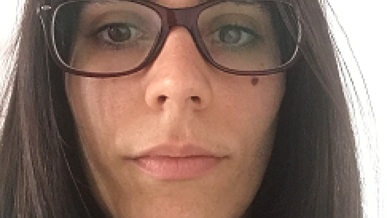 Brunette woman with glasses, neutral expression, plain background.