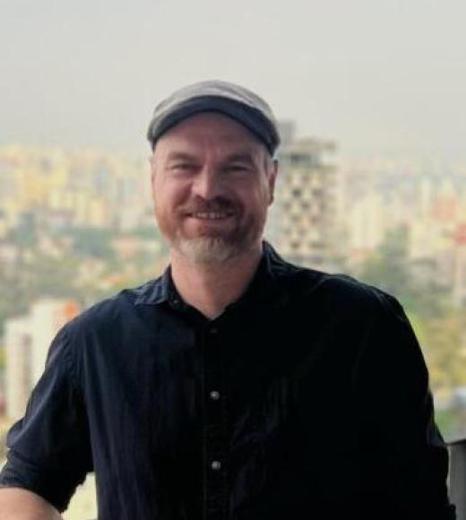 A man wearing a grey had and a black button down shirt. A city skyline in the background