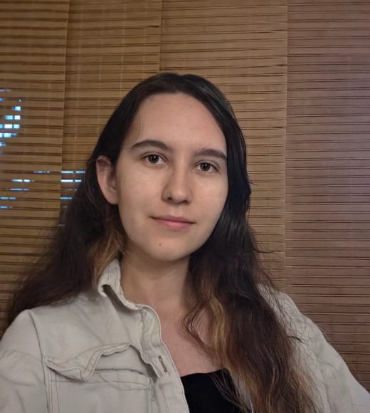 Headshot of young woman with long dark hair, neutral expression, light jacket, wooden blinds