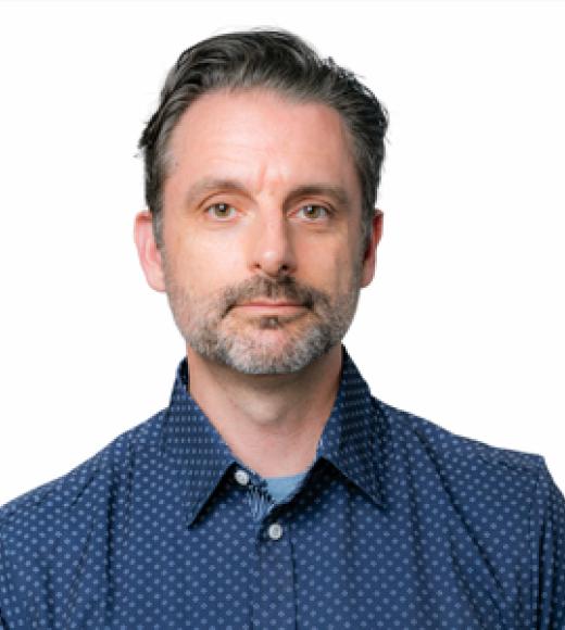 Man with salt-and-pepper hair, short beard, blue shirt, neutral expression, white background.