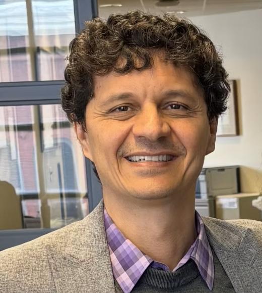 Smiling man with curly dark hair, gray blazer and purple checked shirt in office