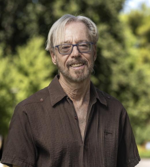 Smiling man with white hair and glasses standing outdoors, blurred green trees, photograph