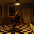 A photo of a man sitting in a room bathed in warm light. He looks at the viewer under one ceiling lamp. A checkered floor.