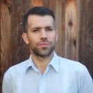 A photo of a man wearing a white shirt in front of a tall wooden fence.
